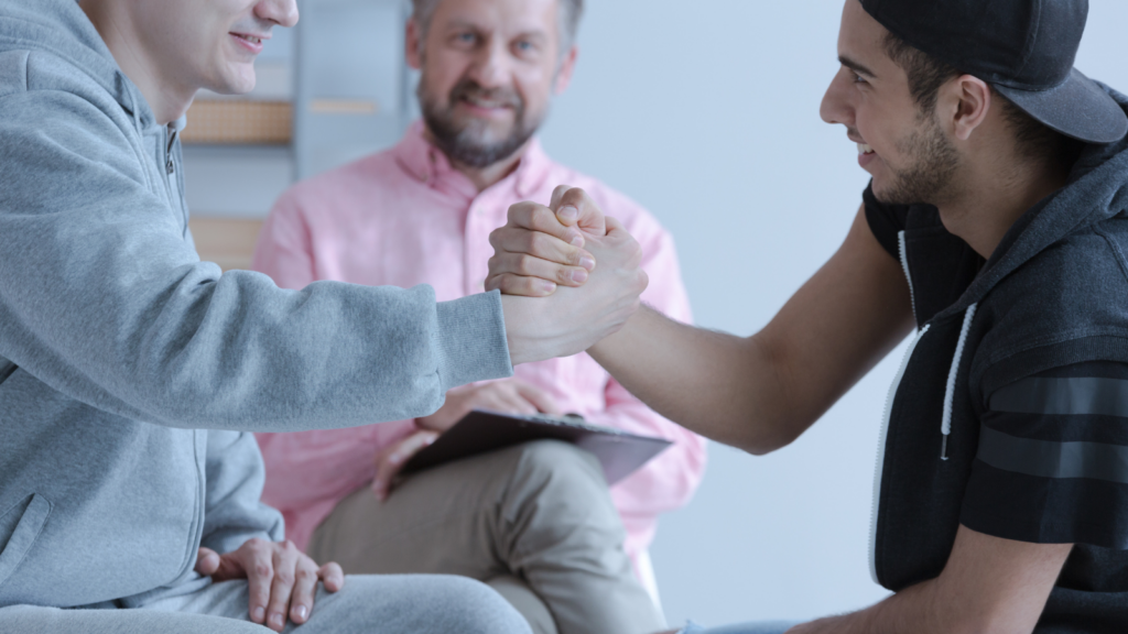 Two young adults clasp hands and smile at each other, while an older man, 40s, looks at them and smiles.