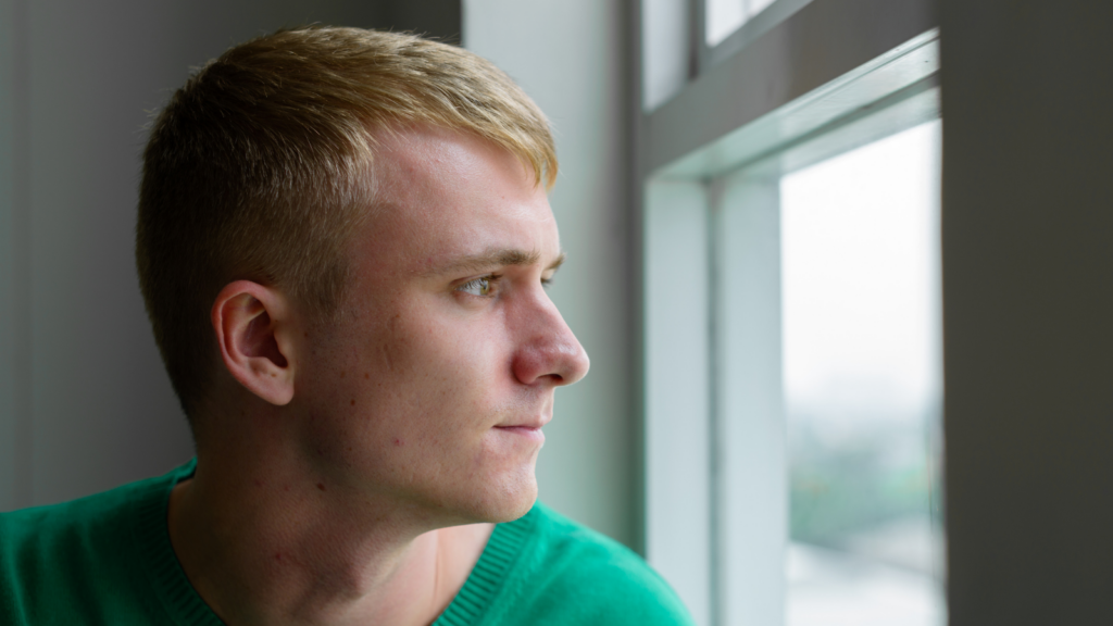 A young white man with blond hair looks out of a window