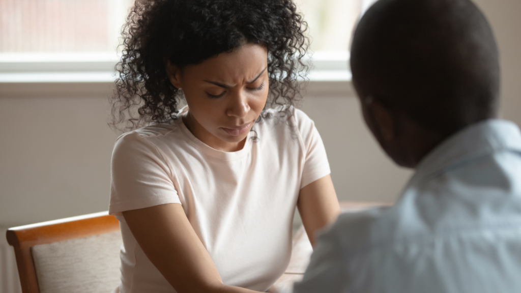 A young mixed race woman looks at the ground and frowns. A young Black man looks at her with his back to the camera.