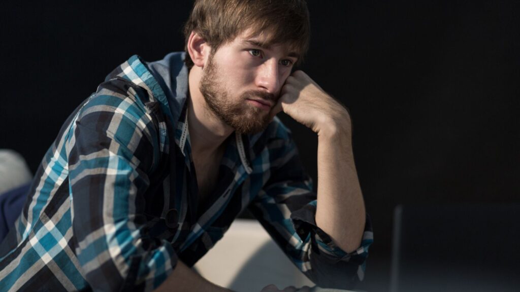 A glum young white man with a beard sits with his head propped up by his hand.