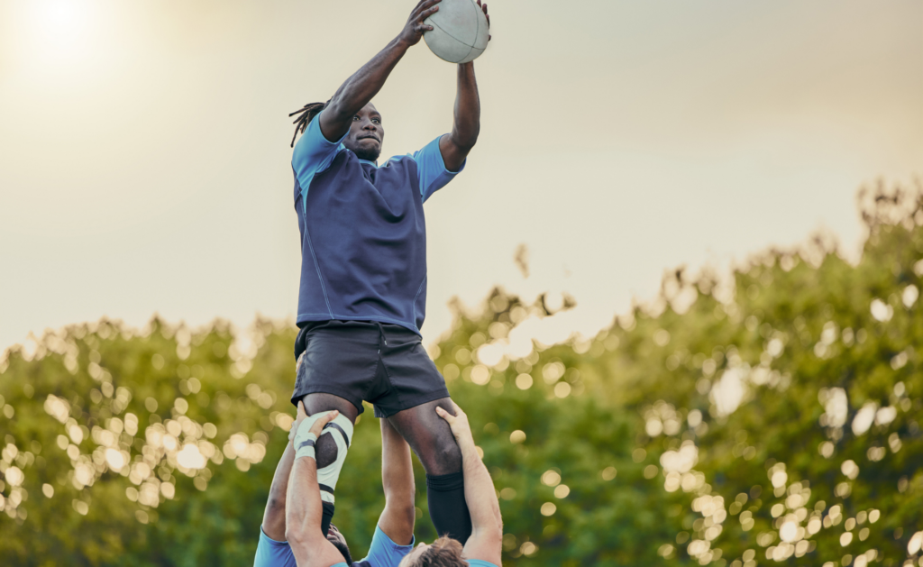 A young Black man catches a rugby ball in the air while held aloft by his team mate