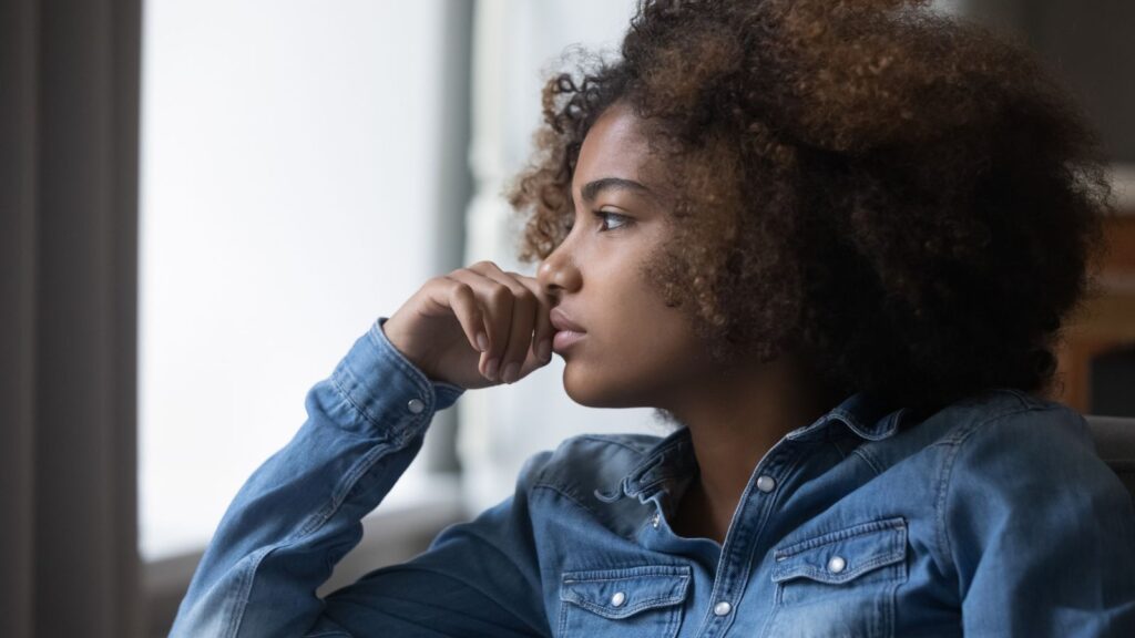 A young Black woman wearing a denim shirt looks thoughtfully out a window